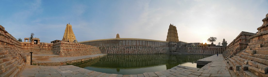 Ancient Hindu temple complex with two stone tower structures flanking a large rectangular water tank surrounded by stone steps and walls under a blue sky at sunset