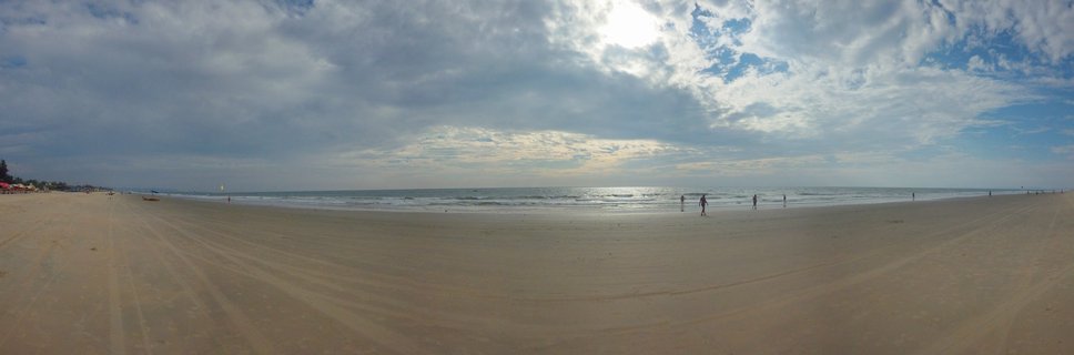 Panoramic photograph of a wide sandy beach in Goa with people walking near the water, ocean waves, and cloudy sky with sun visible through clouds