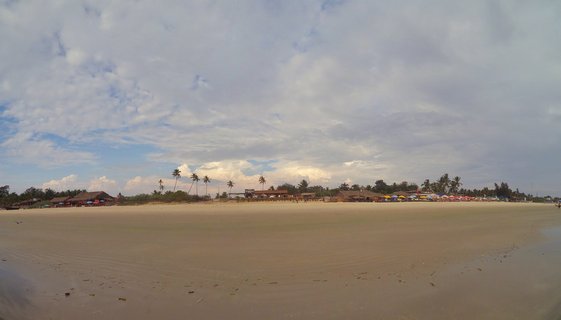 Wide beach in Goa, India showing sand, palm trees, beach shacks with thatched roofs, and colorful umbrellas under cloudy sky