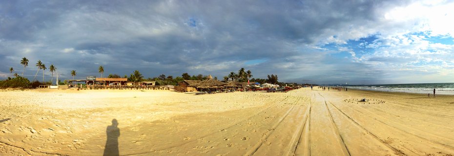 Wide beach panorama showing golden sand with tire tracks, thatched-roof beach huts, palm trees, ocean waves, and people walking under cloudy sky in Goa, India