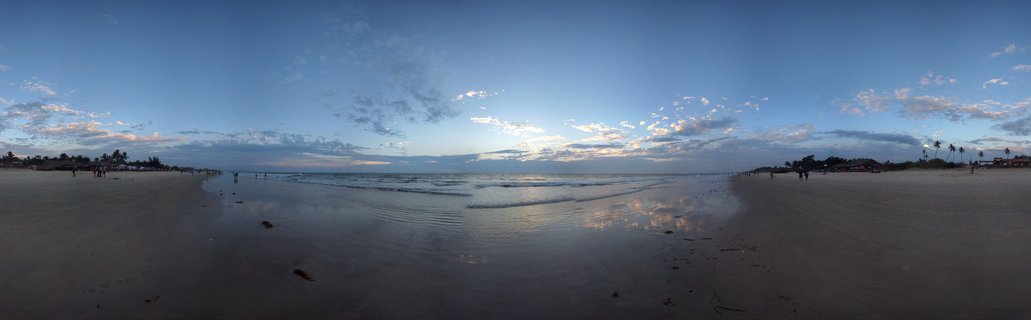 Wide panoramic beach scene in Goa, India showing wet sand reflecting the sky, gentle ocean waves, palm trees along the horizon, and scattered people on the shore during sunset