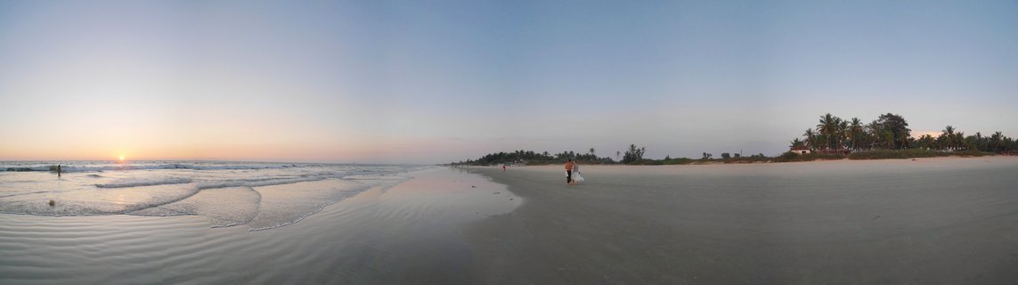 Wide panoramic beach scene at sunset showing ocean waves on the left, two people walking on wet sand, and palm trees along the coastline on the right side in Goa, India