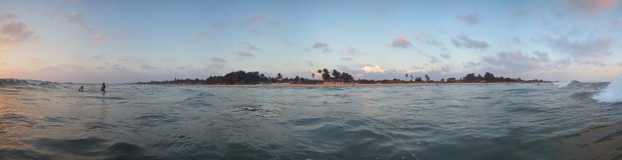 Panoramic beach photograph showing ocean waves in foreground, two people swimming in water, sandy shore with palm trees and buildings in distance under cloudy sunset sky