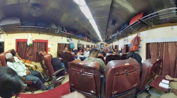 Interior of an Indian Railways train car showing passengers seated and resting in maroon chairs with overhead luggage racks, ceiling fans, and brown curtains along the windows