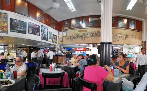 Interior of busy restaurant with people dining at tables with pink and blue tablecloths, murals and framed posters on walls, white columns, and ceiling fans