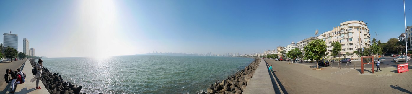 Panoramic view of Marine Drive waterfront promenade in Mumbai with Arabian Sea, Art Deco buildings, people sitting on sea wall, and distant city skyline under bright sun