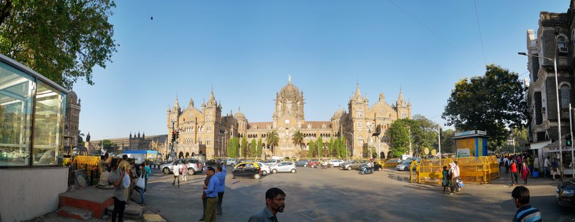 Wide street view of Chhatrapati Shivaji Maharaj Terminus railway station in Mumbai with Victorian Gothic architecture, stone facade, central dome, spires, pedestrians crossing street, cars and yellow-black taxis, traffic barriers, trees, clear blue sky