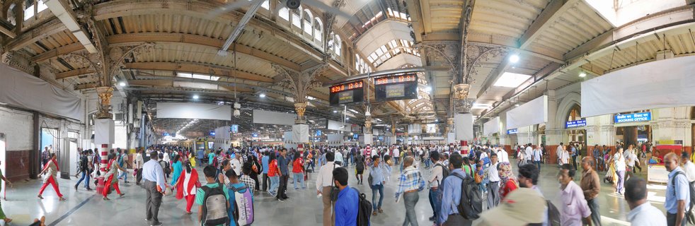 Interior view of a crowded historic railway station with ornate Victorian architecture, vaulted ceilings, cast-iron columns, and hundreds of commuters walking beneath digital train schedule displays