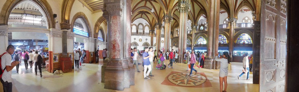 Interior view of Chhatrapati Shivaji Maharaj Terminus railway station showing ornate Victorian Gothic columns, vaulted arched ceilings, stained glass windows, and travelers walking across the marble floor with decorative compass pattern