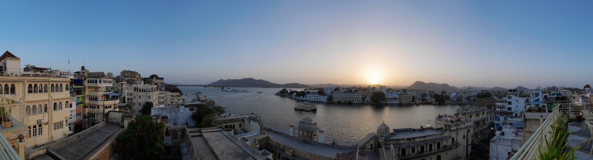 Panoramic sunset view of Lake Pichola in Udaipur showing the Lake Palace on the water surrounded by traditional Rajasthani buildings and distant hills under a clear gradient sky