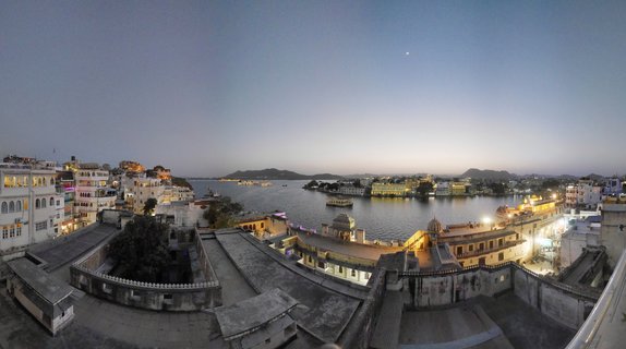 Panoramic view of Lake Pichola in Udaipur at dusk showing the illuminated Lake Palace in the center of the lake, surrounded by traditional white and cream buildings with domed roofs, hills in the background, and a crescent moon in the twilight sky