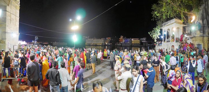 Night photograph showing a large crowd of people on stone steps at a lakeside ghat in Udaipur, India, with an illuminated white palace visible across the water and street lights overhead