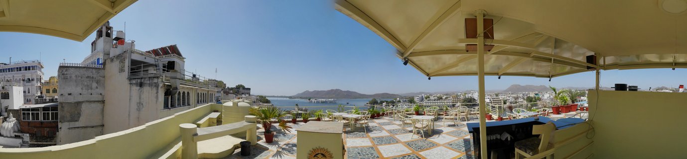 Panoramic view of a rooftop terrace restaurant in Udaipur, India, featuring cream umbrellas and white dining furniture on patterned tile flooring, overlooking Lake Pichola with traditional white Rajasthani buildings and the Aravalli hills visible in the distance under a clear blue sky