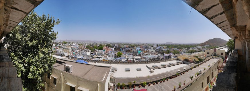 Wide panoramic photograph showing Udaipur city from above with white and cream buildings, flat rooftops, stone architectural columns framing the view, and hills in the distance under clear blue sky
