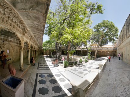 Wide courtyard at City Palace Udaipur with carved stone colonnade on left, patterned marble floor, large green trees in center, stone benches, and visitors walking under bright sunlight