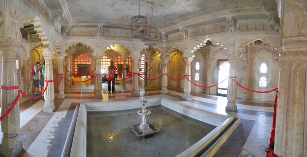 Interior of ornate marble palace hall with carved pillars, arched doorways, central fountain, and tourists walking through the heritage space in Udaipur, India
