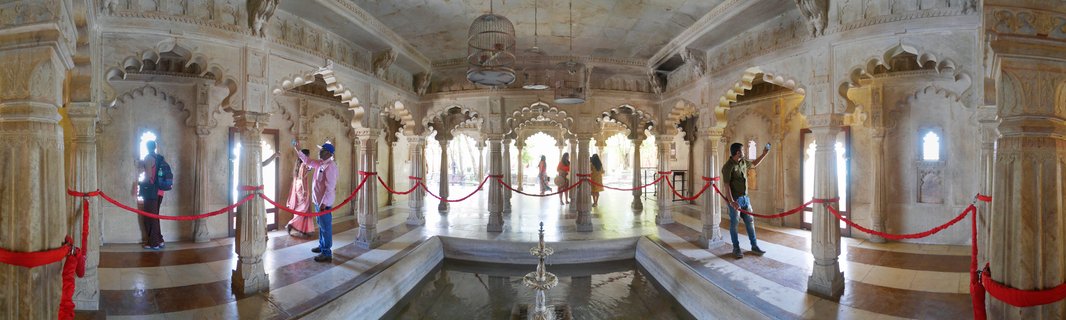 Interior view of ornate palace hall with carved marble columns, arched doorways, central fountain, and visitors walking through the heritage building in Udaipur