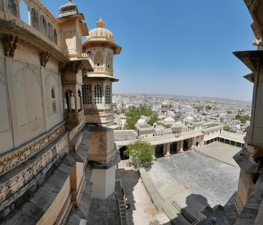 Ornate sandstone palace architecture with domed towers and carved windows overlooking a stone courtyard and Udaipur cityscape under blue sky