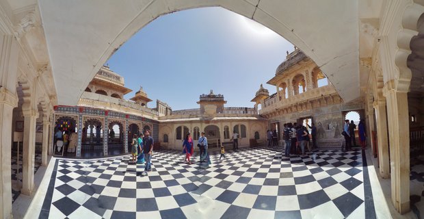 Wide-angle view of tourists walking on a black and white checkered marble courtyard floor inside Udaipur City Palace, with ornate cream-colored Rajasthani architecture, arched colonnades, and domed pavilions under bright blue sky