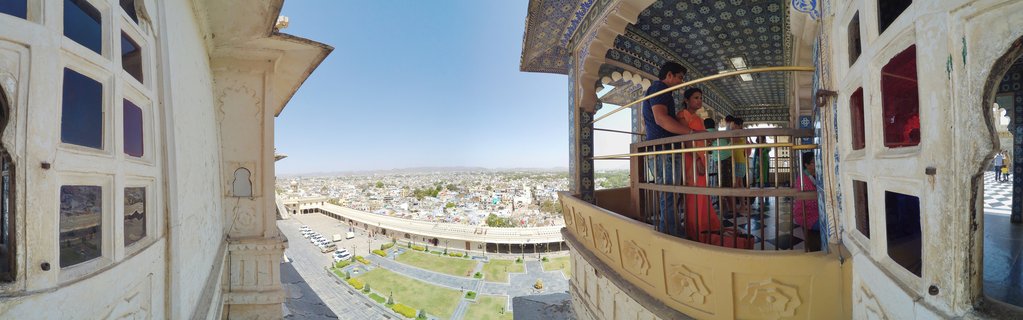 Tourists on decorated palace balcony with blue mosaic ceiling overlooking Udaipur cityscape in Rajasthan, India