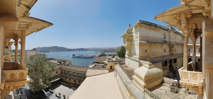 Ornate cream-colored palace balconies and courtyards overlooking blue Lake Pichola with white palace hotel and distant mountains under clear sky in Udaipur, India