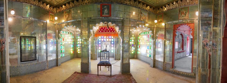 Ornate palace interior with mirror-covered walls, colorful stained glass windows in arches, a wooden throne chair under a red decorative canopy, Persian rug on marble floor, and framed paintings on walls