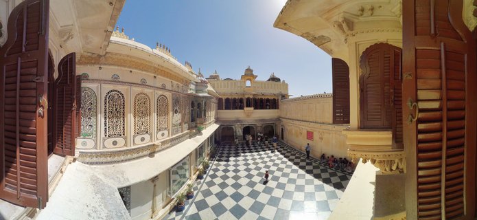 Aerial view of an ornate palace courtyard with black and white checkered marble floor, cream-colored buildings with decorative lattice windows, dark wooden shutters, and tourists walking below under a clear blue sky