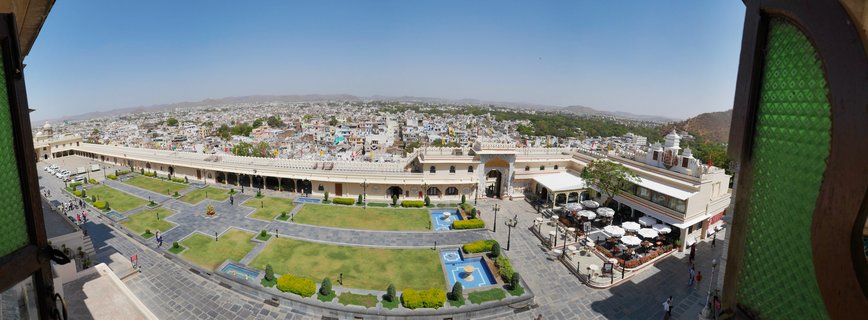 Panoramic view of Udaipur City Palace courtyard with green lawns, blue fountains, and the city skyline with hills in the background under clear blue sky