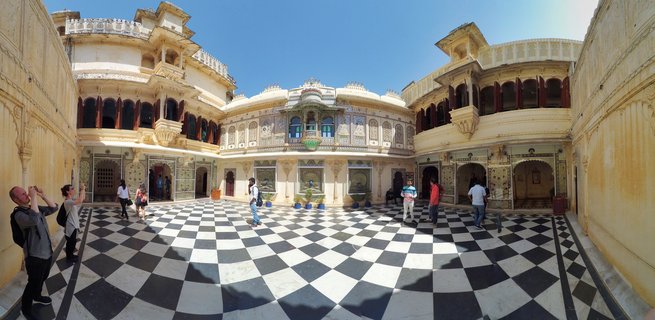 Wide courtyard of Udaipur City Palace with black and white checkered marble floor, cream-colored ornate buildings with balconies and arched windows, tourists walking and taking photographs under blue sky