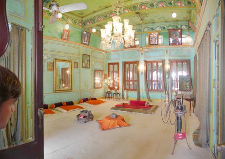 Ornate palace interior room with mint green walls, crystal chandeliers, traditional floor cushions in orange and red, framed paintings, mirrors, and wooden windows in Udaipur, India