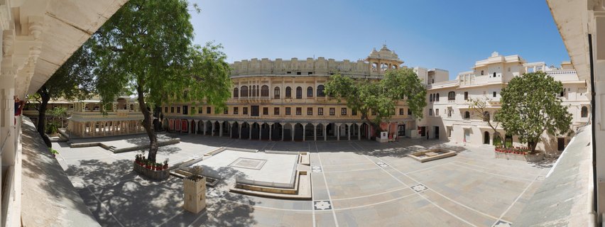 Wide courtyard of City Palace Udaipur with cream-colored historic buildings, arched walkways, large green trees, and geometric stone paving under blue sky