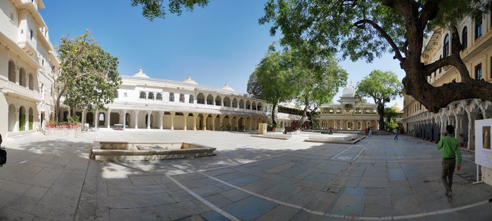 Wide view of City Palace courtyard in Udaipur showing white stone buildings with arched colonnades, large trees, stone pavement, and visitors walking under clear blue sky