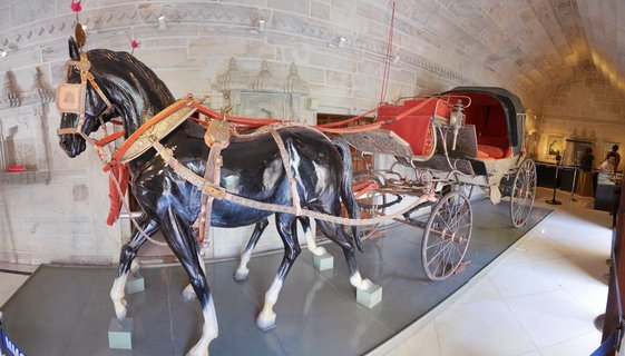 Museum display of a black horse statue with white leg markings harnessed to an ornate royal carriage with red interior, exhibited in a white stone gallery