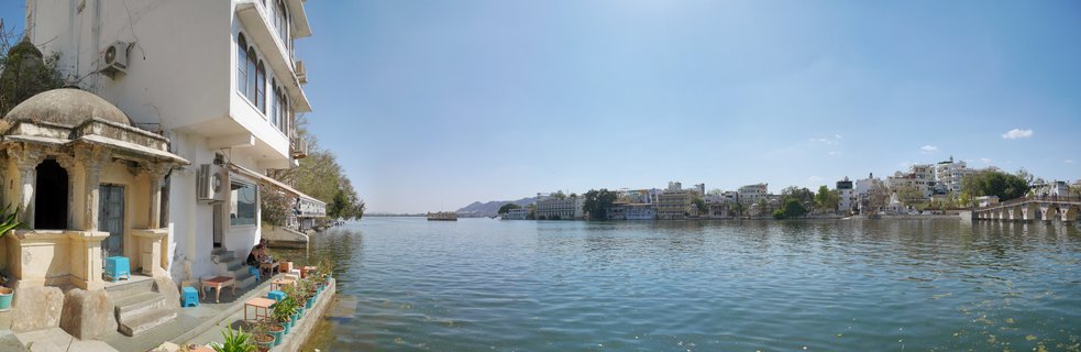 Panoramic view of Lake Pichola in Udaipur showing white Rajasthani buildings with domed pavilions along the waterfront, calm blue-green water, and the Lake Palace in the distance under a clear sky.