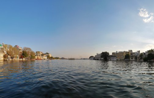 Wide view of Lake Pichola in Udaipur with historic buildings and palaces along both shores under a clear blue sky with sunlight reflecting on the water surface