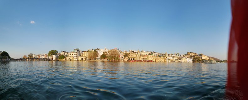 Panoramic view of historic buildings and palaces along Lake Pichola waterfront in Udaipur, India, with blue water in foreground and clear sky above