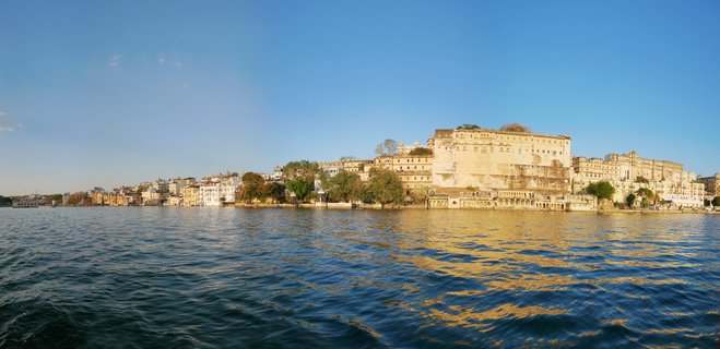 Panoramic view of City Palace Udaipur waterfront with historic buildings along Lake Pichola shoreline under clear blue sky