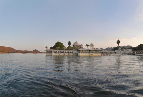 White marble Jag Mandir Palace on an island in Lake Pichola with domes, palm trees, and aravalli hills in the background during golden hour