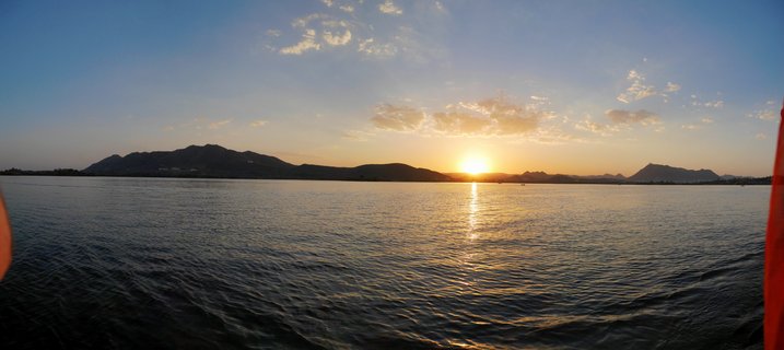 Panoramic sunset photograph showing the sun setting behind silhouetted mountains over a large lake in Udaipur, India, with golden light reflecting on rippling water and scattered clouds in a blue sky