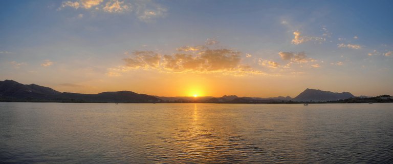 Panoramic sunset view over a lake in Udaipur with the sun setting behind distant mountains, golden light reflecting on water with small boats visible