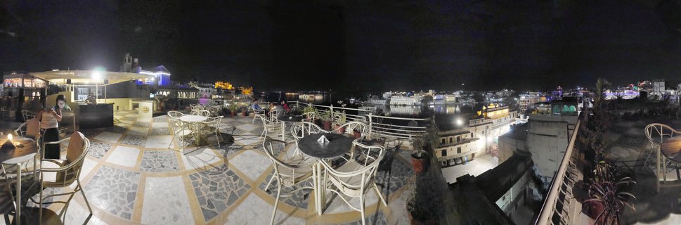 Night view from a rooftop restaurant terrace showing white tables and chairs on patterned tile flooring, with illuminated buildings and Lake Pichola visible in the background in Udaipur, India