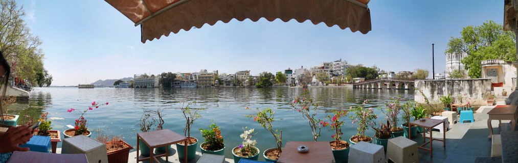 Waterfront cafe terrace with tables and potted flowering plants overlooking Lake Pichola with white Rajasthani buildings and palace architecture on the opposite shore in Udaipur, India