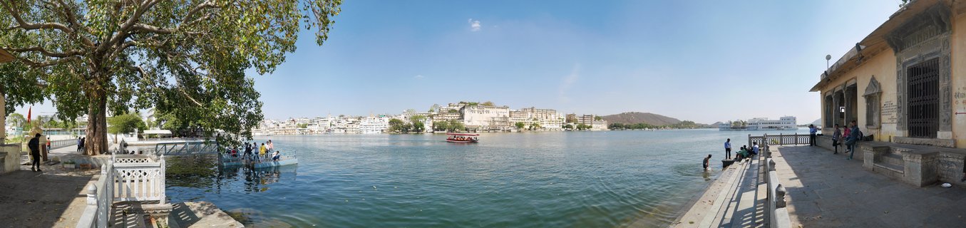 Wide view of Lake Pichola in Udaipur showing white palace buildings across turquoise water with tourist boats and people walking on stone ghats under blue sky