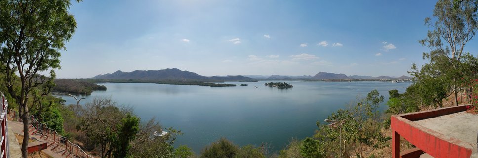 Wide panoramic photograph of Fateh Sagar Lake in Udaipur showing blue water with small islands, distant mountains, trees in foreground, and red viewing structure on the right side