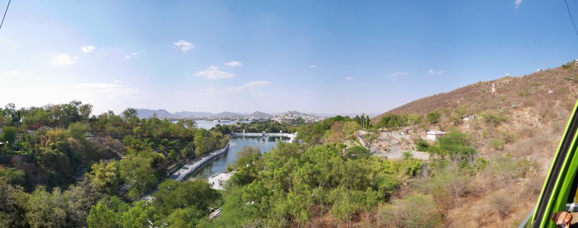 Panoramic aerial photograph showing a lake surrounded by green trees and brown hills under a blue sky with scattered clouds in Udaipur, India