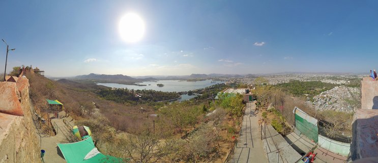 Wide panoramic photograph showing Udaipur city with a large lake in the center, surrounded by dry hills and distant mountains under a bright blue sky with the sun visible