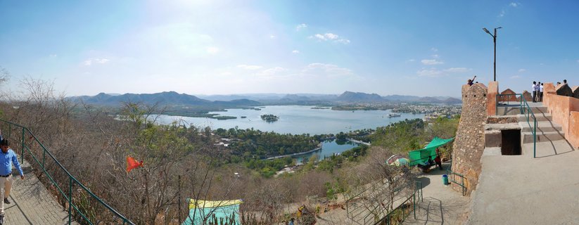Panoramic view from Sajjangarh Fort showing Lake Fateh Sagar with small islands, surrounded by Aravalli hills under clear blue sky. Stone fortification walls and viewing platform visible on right with tourists. Green railings line pathway on left. Orange flags among sparse dry vegetation. Udaipur, Rajasthan, India.