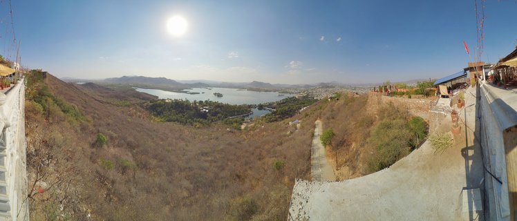 Panoramic landscape showing a large lake surrounded by dry brown hills and distant mountains, with stone walls and viewing structures on both sides, clear blue sky with bright sun overhead