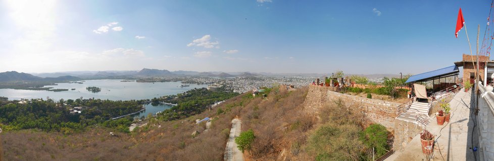 Panoramic aerial view showing Udaipur city with white buildings surrounding Lake Pichola, hills in background, stone fortification walls in foreground with red flag, clear blue sky