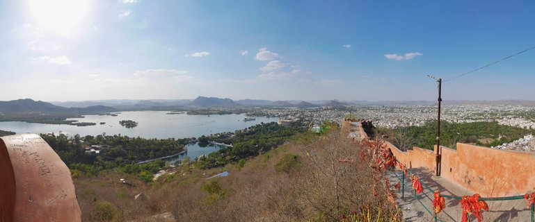 Panoramic photograph showing Lake Pichola with the Lake Palace, the white city of Udaipur, and distant mountains viewed from an elevated fort position with red ceremonial cloths in the foreground